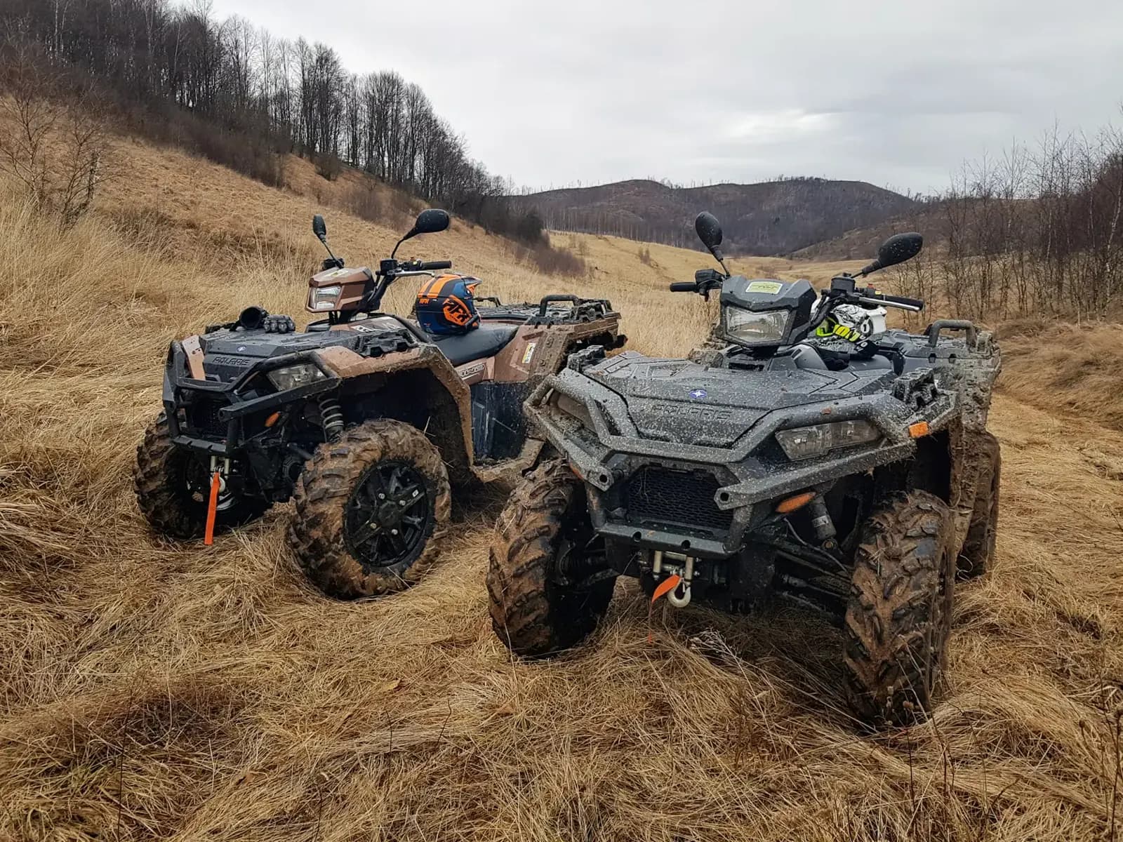 Two mud-covered ATVs parked in tall grass on a hillside trail