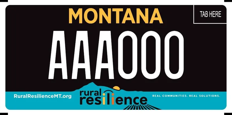 A license plate with yellow, white, and black text. Three fourths of plate black and bottom, one fourth is teal with a mountain peak and sun setting. Bottom says Rural Resilience MT dot org; Rural Resilience; Real Communities; Real Solutions.